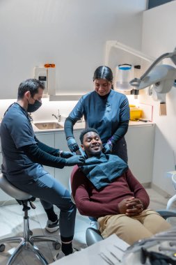 Diş hekimi ve asistanı neşeli bir hastayı diş tedavisi için hazırlıyor. Modern bir diş sağlığı kliniğinde konforunu ve güvenliğini sağlıyor.