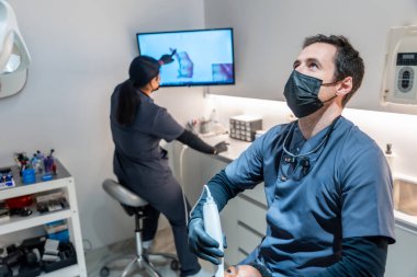 Diş hekimi ultraviyole lamba kullanarak diş beyazlatma prosedürü uyguluyor. Diş hekimi arka planda bir radyografiyi analiz ediyor.