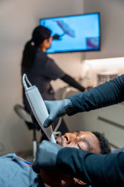 Diş hekimi UV lambası kullanan bir hastaya diş beyazlatma prosedürü uyguluyor, modern bir klinikte profesyonel diş bakımı sergiliyor.