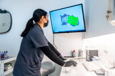 Diş hekimi yüz maskesi ve eldiven takıyor bilgisayar kullanıyor ve diş kliniğindeki bir monitörde 3 boyutlu diş taramasını analiz ediyor.