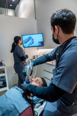 Hastanın dişlerinde diş taraması kullanan diş hekimi ve arka planda 3 boyutlu model görüntülendi.