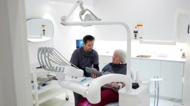 Profesyonel dişçi, dijital tablet kullanarak diş tedavisi detaylarını açıklarken, modern diş sağlığı kliniğindeki yaşlı kadın hastaya da danışıyor.