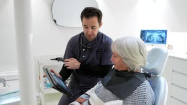 Dişçi yaşlı kadınlara teşhis koyuyor, modern diş sağlığı kliniğinde dijital tablet üzerinde sonuçlar gösteriyor.