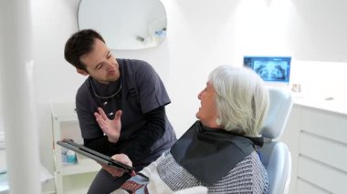 Modern klinik ortamında tam diş muayenesi ve muayenesi sırasında dijital tablet kullanan yaşlı kadına tıbbi teşhisi açıklayan deneyimli bir dişçi.