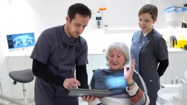 Modern klinikte kapsamlı oral sağlık danışmanlığı sırasında kıdemli hastaya dijital tablet üzerindeki diş röntgenini açıklayan deneyimli bir diş hekimi