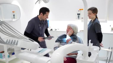 Dişçi ve asistan, modern bir diş kliniğinde tablet kullanan kıdemli bir hastayla tedavi planını tartışıyor.