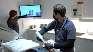 Diş hekimi yaşlı hastaya diş beyazlatma işlemi için ultraviyole lamba kullanırken hemşire de ekrandaki röntgene bakarak ona yardım ediyor.