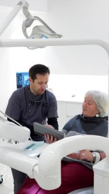 Dişçi, modern diş kliniğindeki tablet ekranı göstererek yaşlı kadınlara teşhis koyuyor.
