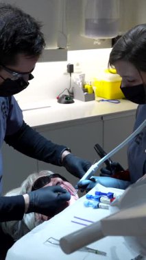 Diş hekimi ve asistanı dişçi kliniğinde kıdemli bir kadın hastaya diş beyazlatma prosedürü uyguluyor.