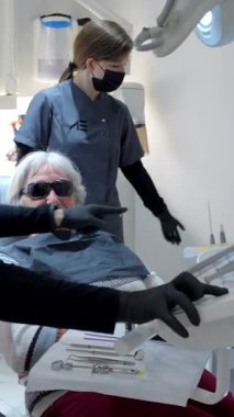 Yaşlı bir kadın diş tedavisi görüyor, koruyucu gözlük takıyor ve ameliyat sırasında ağzını çalkaluyor.