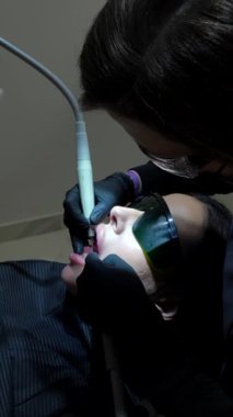 Profesyonel dişçi diş beyazlatma prosedürünü özel diş ekipmanlarıyla gerçekleştiriyor, klinik ortamdaki kadın hastaları dikkatlice tedavi ediyor.