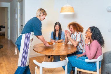 Modern bir dairede bir kadın onlara yemek servisi yaparken mutlu çok ırklı arkadaşlar el çırpıyor.