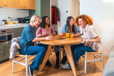 Dört farklı oda arkadaşı yuvarlak bir mutfak masasında oturup gülüyor ve kahvaltıdan keyif alıyorlar.