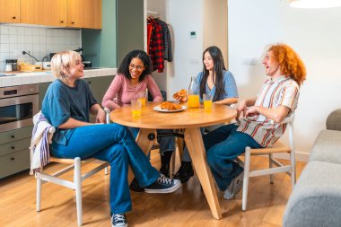 Dört farklı genç insan yuvarlak bir masada oturuyor, birlikte kahvaltı ederken gülüyor ve sohbet ediyorlar.