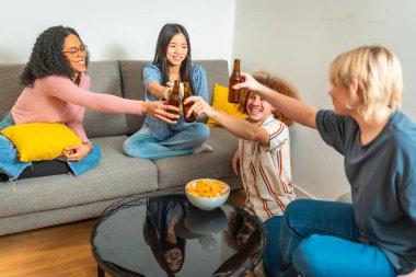 Bir grup etnik kökenli genç kanepede oturup cips yerken bira şişeleriyle kadeh kaldırıyor ve eğleniyorlar.