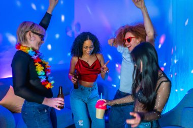 Neşeli çoklu etnik arkadaşlar birlikte dans ediyor ve mavi ve pembe ışıklandırmalı bir disko partisinde içki içiyorlar.