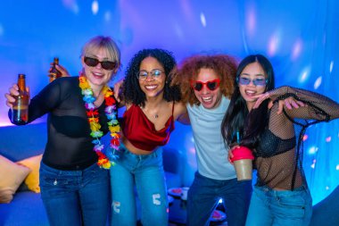 Dört farklı arkadaş renkli ışıklandırma ve içeceklerle coşkulu bir ev partisinde eğleniyor, sarılıyor ve gülümsüyor.