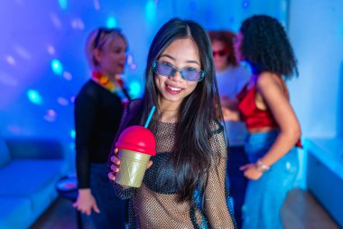 Asyalı kadın elinde renkli bir içecek tutuyor, gülümsüyor ve arkadaşlarıyla bir gece kulübü partisinin canlı atmosferinden zevk alıyor.