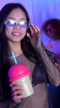Dans ederken renkli milkshake tutan genç bir kadın, müzik ve bulaşıcı enerji ile canlı parti atmosferini deneyimliyor.