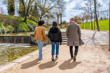 Güneşli parktaki küçük şelaleli gölün yanındaki yolda kameradan uzaklaşan üç arkadaş.