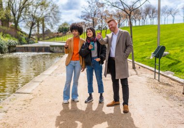 Üç arkadaş parkta gölet kenarında yürüyorlar güneşli bir günün tadını çıkarıyorlar ve dikkatlerini çeken bir şeye işaret ediyorlar.