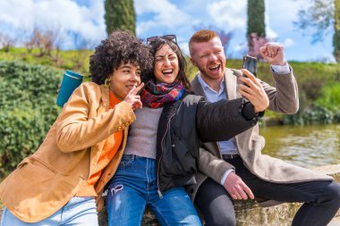 Üç neşeli çoklu etnik arkadaş gölün yanındaki bir duvarda otururken selfie çekiyorlar.