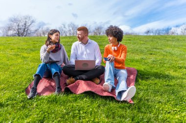Üç genç girişimci açık havada çalışıyor, teknolojik cihazlar kullanıyorlar, güneşli bir bahar gününün tadını çıkarıyorlar.