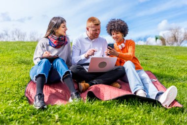 Üç üniversite öğrencisi bir parkta battaniyenin üzerinde otururken dizüstü bilgisayar ve akıllı telefon kullanarak bir projede işbirliği yapıyorlar.