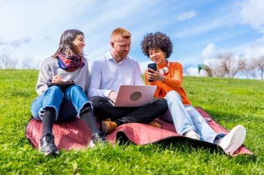 Üç üniversite öğrencisi bir dizüstü bilgisayar, tablet ve telefon kullanarak açık havada bir projede çalışıyor. Bahar havasının tadını çıkarıyorlar.