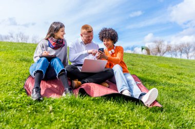 Üç öğrenci çimenlerde dizüstü bilgisayar ve akıllı telefon kullanarak birlikte parkta ders çalışmaktan zevk alıyorlar.