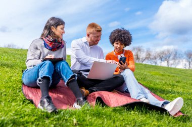 Üç öğrenci bir projede işbirliği yapıyor, dizüstü bilgisayar, tablet ve akıllı telefon kullanıyorlar, parktaki güneşli havanın tadını çıkarıyorlar.