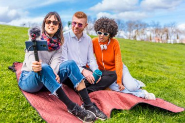 Üç içerikli yaratıcı, parktaki bir video blogunu dış mikrofonlu akıllı bir telefonla kaydediyor ve battaniyenin üzerinde oturuyor.