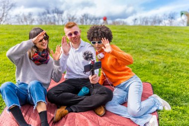 Güneşli bir parkta video blogu çekerken üç içerikli yaratıcı kameraya neşeyle el sallıyor ve birlikte vakit geçiriyorlar.