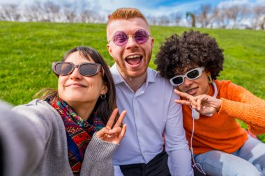 Üç mutlu çoklu etnik arkadaş yeşil bir parkta güneşli bir günün tadını çıkarırken barış işareti yaparken selfie çekiyorlar.
