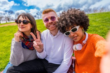Üç farklı genç arkadaş parkta bir battaniyenin üzerinde oturmuş selfie çekiyorlar ve barış işareti gösteriyorlar.