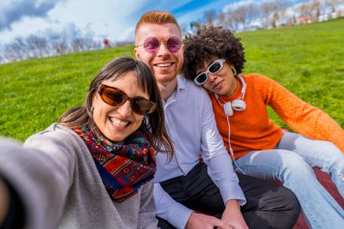 Üç neşeli arkadaş parkta selfie çekiyorlar, güneşli bir bahar gününün tadını çıkarıyorlar.