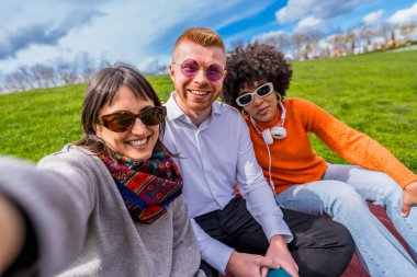 Üç neşeli çoklu etnik arkadaş parkta selfie çekiyorlar, güneşli bir günün tadını çıkarıyorlar.