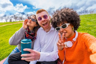 Üç neşeli çoklu etnik arkadaş bir parkta yeniden kullanılabilir kahve fincanıyla fotoğraf çektirirken komik surat ifadeleri takınıp el kol hareketi yapıyorlar.