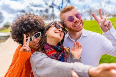 Üç mutlu çoklu etnik arkadaş güneş gözlüğü takıyor. Parkta selfie çekerken barış işareti yapıyorlar.