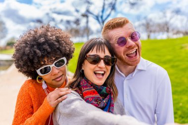 Üç neşeli çoklu etnik arkadaş parkta selfie çekiyorlar, birlikte gülüyorlar ve güneşli güzel bir günün tadını çıkarıyorlar.