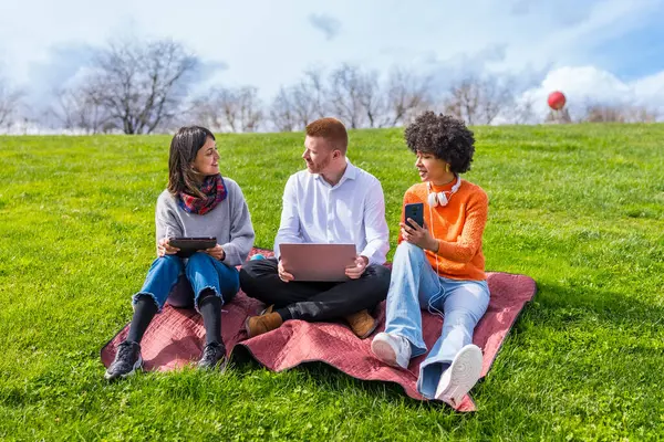 Üç üniversite öğrencisi parktaki bir battaniyede oturup bahar gününün tadını çıkarırken elektronik aletler kullanıyorlar.