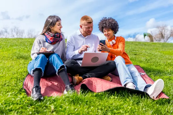 Üç üniversite öğrencisi bir parkta battaniyenin üzerinde otururken dizüstü bilgisayar ve akıllı telefon kullanarak bir projede işbirliği yapıyorlar.
