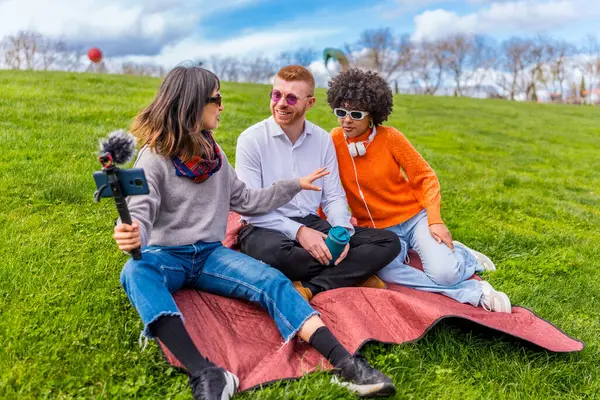 Üç içerik oluşturucu, video yayınları için bir parkta bir röportaj kaydediyor. Selfie çubuğunda akıllı telefon ve harici bir mikrofon kullanıyorlar.