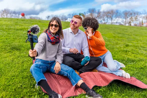 Üç içerik yaratıcısı parkta battaniyeye oturmuş, video blogunu dış mikrofonlu akıllı telefon kullanarak çekiyor, güneşli günün tadını çıkarıyor ve eğleniyorlar.