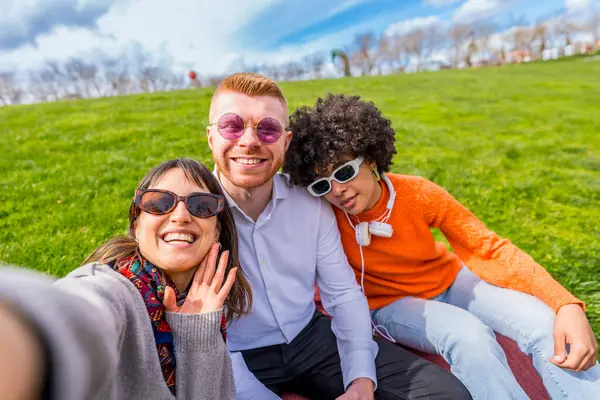 Üç çok ırklı arkadaş parkta çimenlerde oturmuş selfie çekiyor ve kameraya el sallıyorlar.