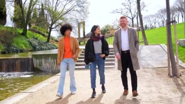 Arkadaşlar birlikte yürüyor, güneşli park manzarasının tadını çıkarıyor, sakin su özelliklerinin yakınındaki yeşil patikada gezinirken canlı sohbetler yapıyorlar.