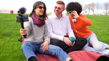 Genç içerik yaratıcıları parkta bir battaniyeye oturmuş, mikrofonlu bir akıllı telefon kullanarak bir video blogu kaydediyorlar.