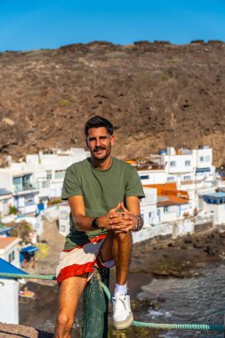 Tahta bir direkte oturan genç adam Tufia 'nın renkli evlerinin, Büyük Kanarya' nın sahil köyünün, kanarya adalarının ve İspanya 'nın manzarasının tadını çıkarıyor.