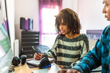 Evden çalışan iki programcı, bilgisayar ekranına bakarken not defterine notlar yazıyorlar.