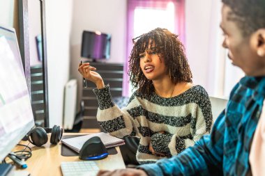 Bir projede işbirliği yapan iki web tasarımcısı, rahat bir ofis ortamında bilgisayar üzerinde çalışırken fikirleri ve çözümleri tartışıyorlar.